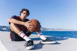 Foto gratuita adolescente con pallacanestro che si siede vicino al mare