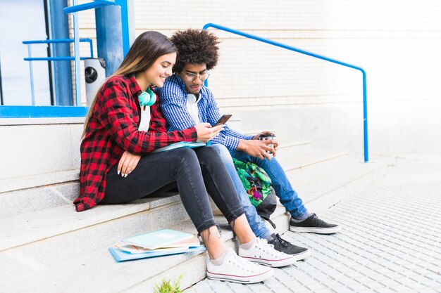 Студент подростковой пары сидя на белой лестнице используя мобильный телефон
