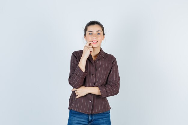 Teenage girl biting finger in brown striped shirt and looking forgetful , front view