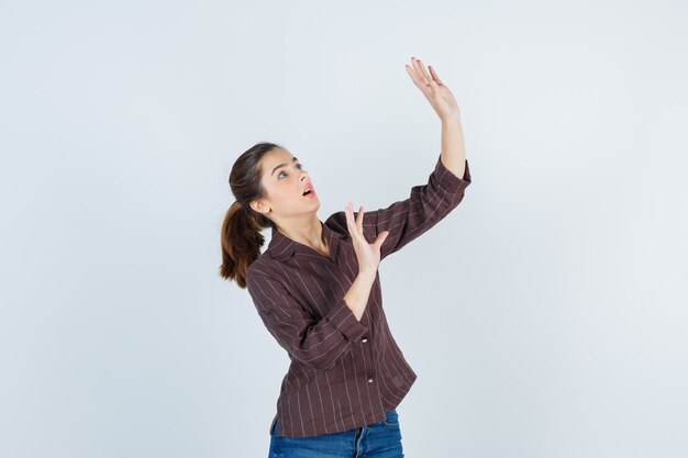 Teenage girl in brown striped shirt showing stop gesture as if trying to defend herself and looking scared , front view.