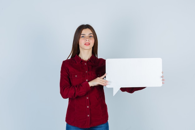 Free photo teenage girl in burgundy shirt holding speech bubble and looking confident , front view.