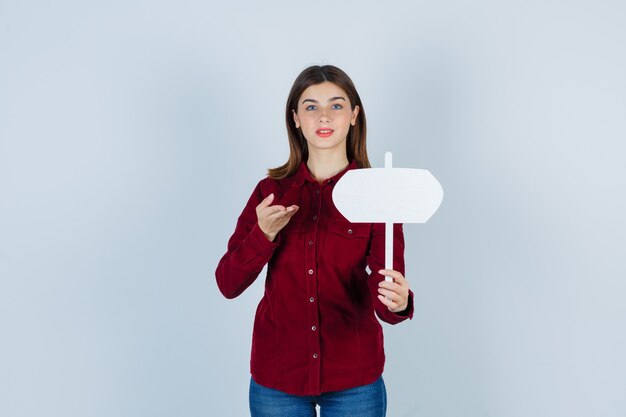 Teenage girl in burgundy shirt showing paper sign and looking hesitative , front view.