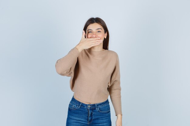 Teenage girl covering mouth with hand in sweater, jeans and looking jovial. front view.