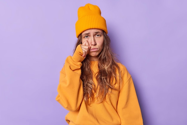 Teenage girl faces problems sobs and wipes tears stands very upset wears casual orange jumper and hat expresses negative emotions isolated on purple feels heartbroken. stress anxiety