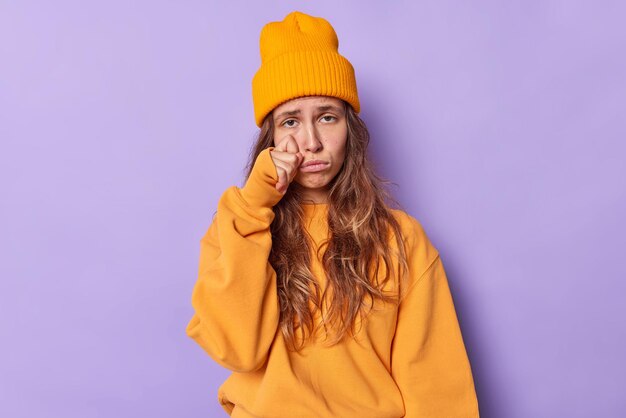 Teenage girl faces problems sobs and wipes tears stands very upset wears casual orange jumper and hat expresses negative emotions isolated on purple feels heartbroken. Stress anxiety