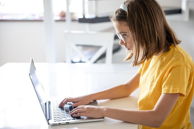 안경을 쓴 십대 소녀 가 집 에서 테이블 에 앉아 노트북 을 사용 하고 있다