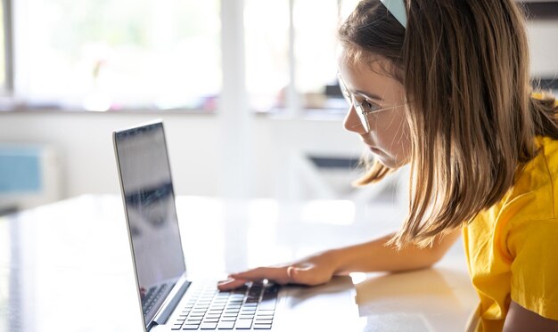 A teenage girl in glasses uses a laptop while sitting at a table at home