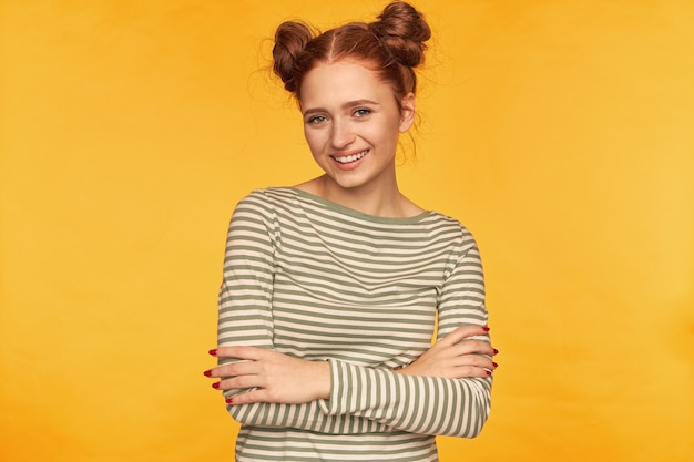 Teenage girl, happy looking red hair woman with two buns. wearing striped sweater and holds hands crossed on a chest. smiling watching isolated over yellow wall