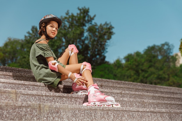Девочка-подросток в шлеме учится кататься на роликовых коньках, держа баланс или кататься на роликах, и крутиться на улице города в солнечный летний день