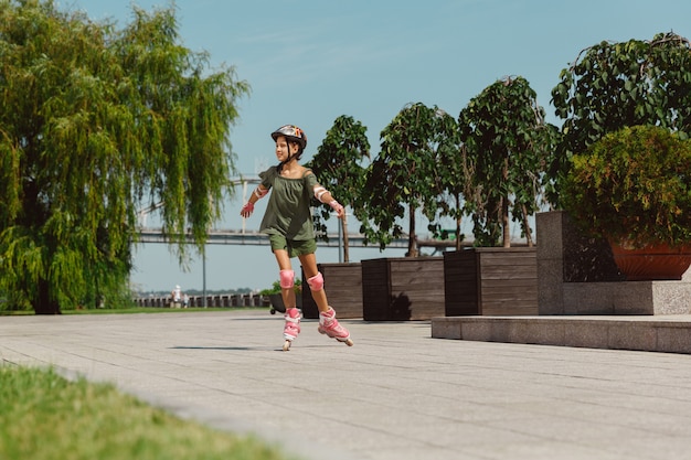 헬멧에 십 대 소녀는 화창한 여름 날에 균형 또는 롤러 블레이드를 들고 롤러 스케이트를 타고 도시의 거리에서 회전하는 법을 배웁니다.