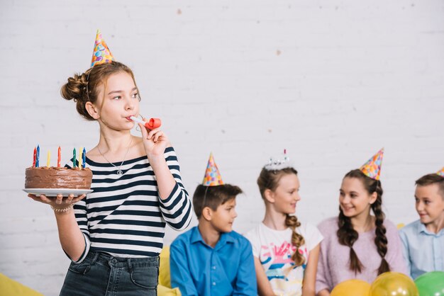 Teenage girl holding birthday cake in hand blowing party horn standing in front of her friends sitting together