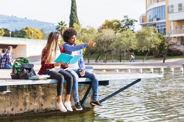 Девочка-подросток держит книгу в руке, глядя на своего парня, показывая что-то возле озера