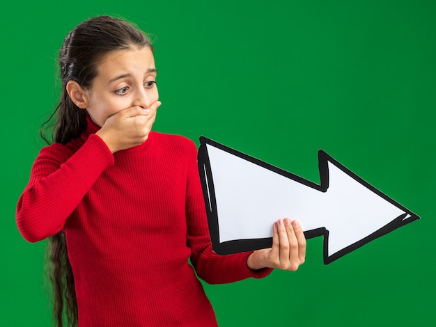 Teenage girl holding and looking at arrow mark pointing at side keeping hand on mouth isolated on green wall