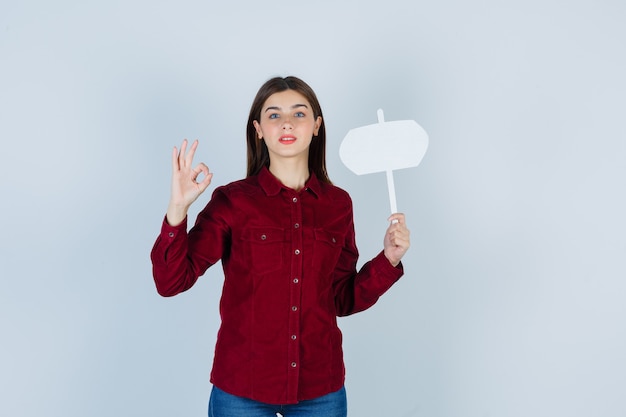 Free photo teenage girl holding paper sign, showing ok gesture in burgundy shirt and looking pleased. front view.