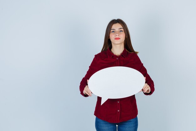 Teenage girl holding speech bubble in burgundy shirt, jeans and looking confident. front view.