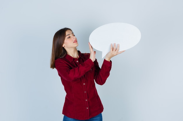 Teenage girl holding speech bubble in burgundy shirt and looking confident. front view.