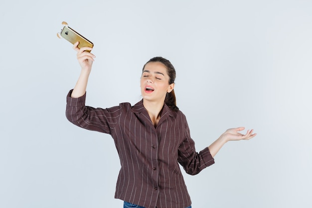Девочка-подросток в коричневой полосатой рубашке держит мобильный телефон, разводит ладонь в сторону, закрывает глаза и выглядит удивленно, вид спереди.