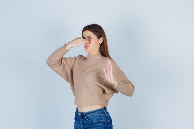 Бесплатное фото Девочка-подросток в свитере, в джинсах, пахнущих чем-то отвратительным, щиплет нос, показывая знак остановки и недовольный вид, вид спереди.