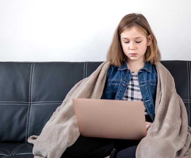 Девушка-подросток сидит на диване с ноутбуком, завернутым в плед
