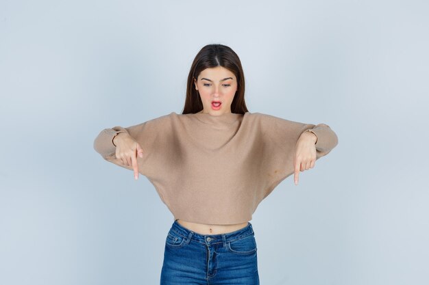 Teenage girl pointing down in sweater, jeans and looking amazed. front view.