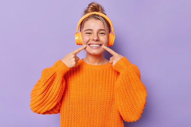 Teenage girl points at beaming smile wears knitted orange sweater listens music via headphones has combed hair isolated over purple background. Look at my perfect teeth. People and hobby concept