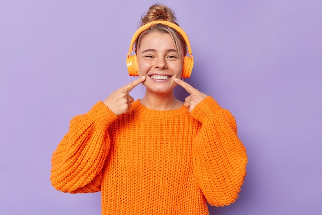Девочка-подросток указывает на сияющую улыбку, носит вязаный оранжевый свитер, слушает музыку через наушники, расчесывает волосы, изолированные на фиолетовом фоне. Посмотрите на мои идеальные зубы. Концепция людей и хобби