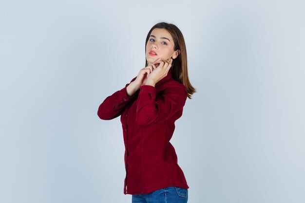 Teenage girl posing while touching her jaw in burgundy shirt and looking charming , front view.