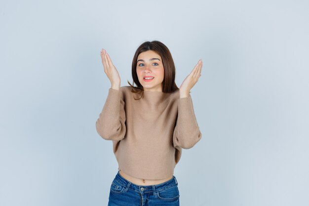 Teenage girl raising hands in clueless manner in sweater, jeans and looking jolly , front view.