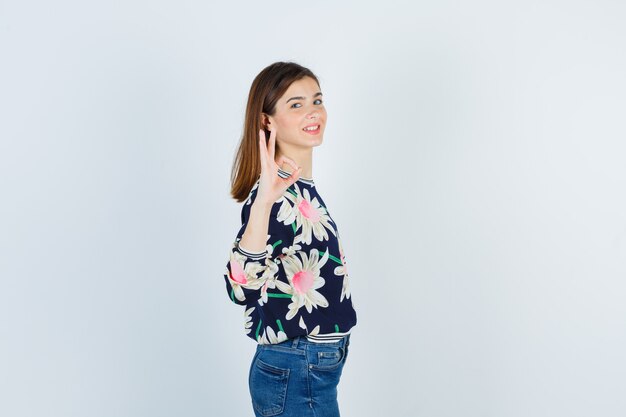 Teenage girl showing ok gesture in blouse, jeans and looking confident. front view.