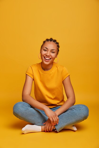 Teenage girl smiles broadly, sits upbeat and relaxed on floor, wears casual clothes, feels positive, enjoys spare time, crosses legs isolated on yellow  wall has fun at home. Emotions, lifestyle