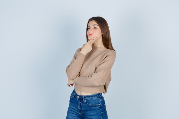 Teenage girl in sweater, jeans keeping fingers on jaw and looking thoughtful , front view.