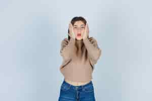 Free photo teenage girl in sweater, jeans keeping hands on cheeks and looking anxious , front view.