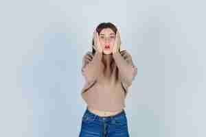 Free photo teenage girl in sweater, jeans keeping hands on cheeks and looking anxious , front view.