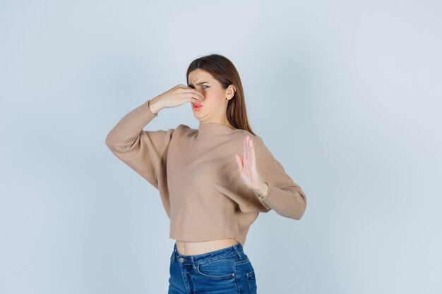 Teenage girl in sweater, jeans smelling something disgusting, pinching nose, showing stop sign and looking displeased , front view.