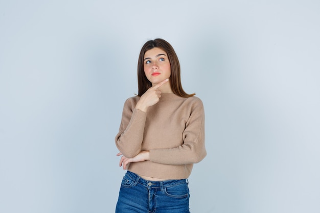 Adolescente in maglione, jeans che toccano la mascella con il dito, alzando lo sguardo e guardando pensieroso, vista frontale.