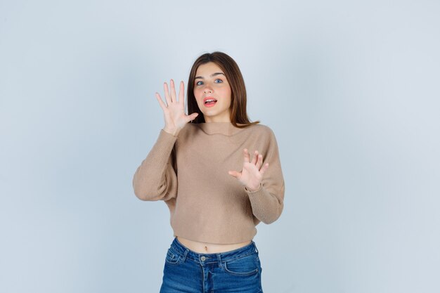 Teenage girl trying to block herself with hands in sweater, jeans and looking scared , front view.