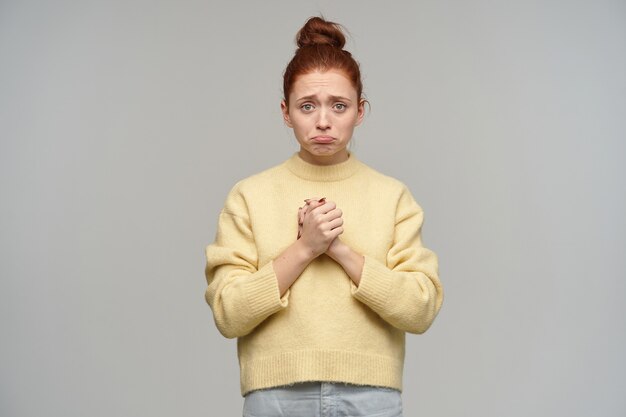 Teenage girl, upset looking woman with ginger hair gathered in a bun. wearing pastel yellow sweater and jeans. keep arms together, feels sorry.  isolated over grey wall