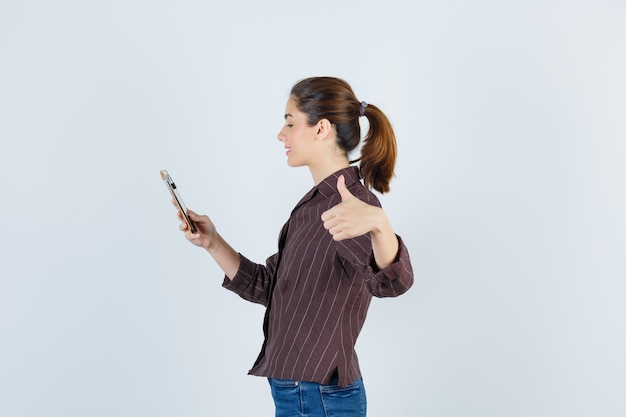 Teenage girl using smartphone, showing thumb up in brown striped shirt and looking pleased. .