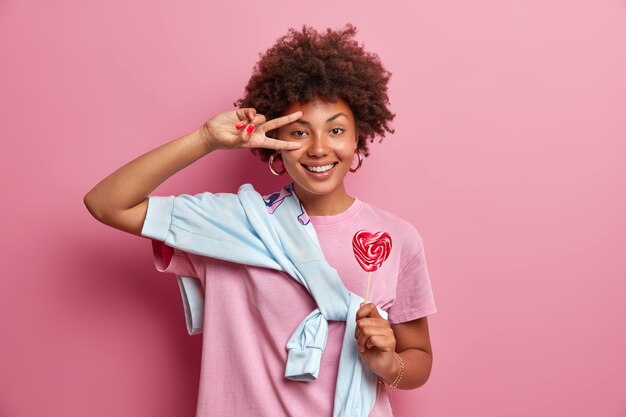 Teenage girl with curly hair makes victory gesture over eye, has happy expression, holds appetizing lollipop on stick, dressed casually, poses 