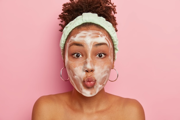 Teenage girl with dark skin has problematic skin, applies foam to wetter face, keeps lips folded