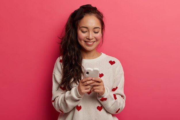 Девочка-подросток с нежной улыбкой набирает сообщения на современном мобильном телефоне, общается в чате, читает сообщения в социальных сетях, страдает технологической зависимостью.