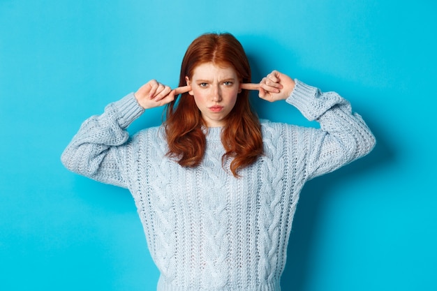 Teenage redhead girl unwilling to listen, shut ears and frowning angry, staring at camera offended, sulking against blue background