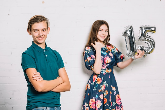 Teenage smiling girl holding numeral 15 foil balloon pointing finger toward the birthday boy