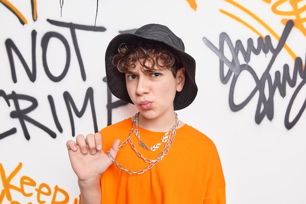 10代のストリートアーティストは帽子オレンジ色のTシャツを着ており、落書きの壁に対してチェーンポーズは不満の表現を持っています