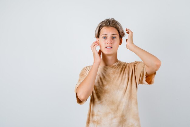 Teenager boy in t-shirt keeping hands near head and looking troubled , front view.