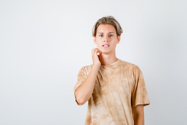 Teenager boy in t-shirt touching neck and looking troubled , front view.