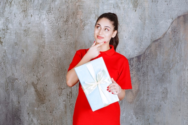 Teenager girl holding a blue gift box wrapped with white ribbon and dreaming or having a good idea