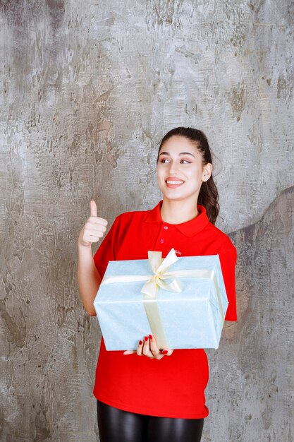 Teenager girl holding a blue gift box wrapped with white ribbon and showing enjoyment hand sign. 