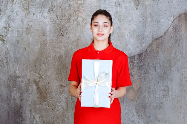 Девушка-подросток держит голубую подарочную коробку, обернутую белой лентой.