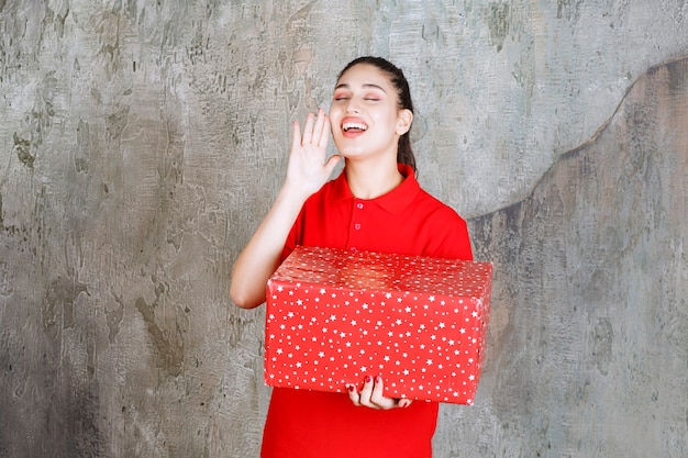 Teenager girl holding a red gift box with white dots on it and calling for someone